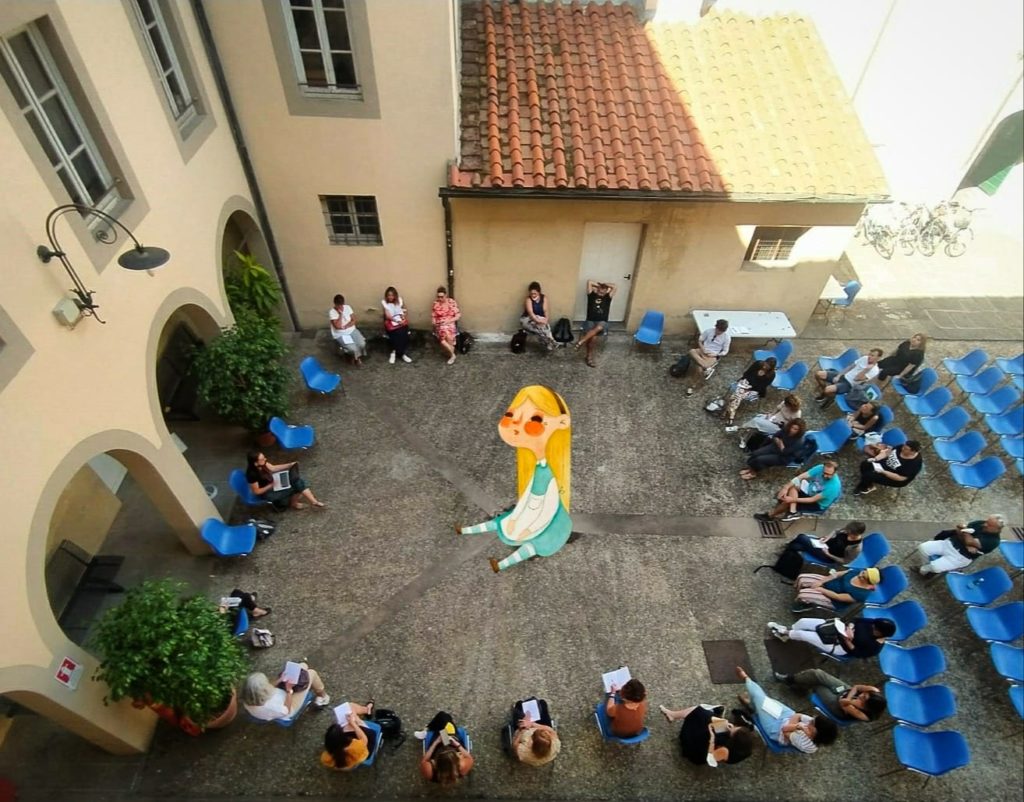 Un grande cerchio di lettrici e lettori seduti in un cortile, visto dall'alto